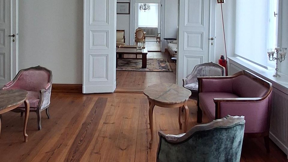 Heller, klassischer Salon in einem Altbau mit hohen Decken, Stuckgesims und großem Kristalllüster über breitem Holzdielenboden. Im Raum stehen mehrere Polstersessel und kleine Beistelltische; doppelflügelige Türen öffnen den Blick in weitere, hintereinanderliegende Zimmer mit großen Fenstern.