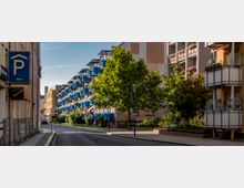 Wohnstraße mit mehrstöckigen Wohngebäuden, die durch zahlreiche blau gestrichene Balkone geprägt sind. Auf der linken Straßenseite befindet sich ein Schild mit Parkhinweis, während Bäume und Blumen auf der rechten Seite den Bereich begrünen.
