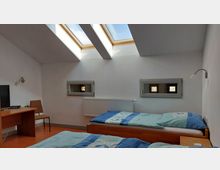 Dachgeschosszimmer mit zwei Einzelbetten, hellgrauen Wänden und einem Dachfenster, das Tageslicht einfällt lässt. Es gibt einen Schreibtisch mit Stuhl, eine Wandheizung unter zwei kleinen rechteckigen Fenstern und eine Lampe an jeder Bettseite.