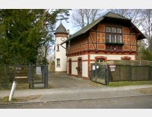 Zweigeschossiges historisches Gebäude mit Fachwerkfassade und Ziegeltönen, daneben ein angeschlossener runder Turm mit spitzem Dach. Im Vordergrund ein Metalltor mit Hausnummer „11-15“ und ein Gehweg, umgeben von Bäumen und einer umzäunten Einfahrt.