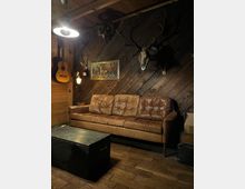 Rustikaler Aufenthaltsraum mit schräg vertäfelten Holzwänden und Holzboden, vor einer braunen Ledercouch und einem schwarzen Kistentisch. An der Wand hängen Jagdtrophäen (Geweihe und Schädel), ein Waldgemälde sowie eine Gitarre; warme Lampen beleuchten den Raum.