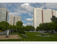 Mehrere Hochhäuser im Hintergrund mit weißer Fassade und roten Balkonen, umgeben von grünen Bäumen. Im Vordergrund ein kleiner Park mit Rasen, einer Rutsche auf einem Sandspielplatz, einer Tischtennisplatte und wenigen Sitzbänken.