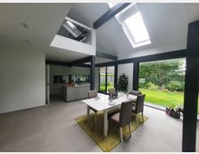 Ein modernes Esszimmer mit offener Küche, großen Glasfenstern und Blick auf einen grünen Garten. Die Einrichtung umfasst einen weißen Esstisch mit vier Stühlen auf einem gelben Teppich und eine schlichte Küchenzeile im Hintergrund.