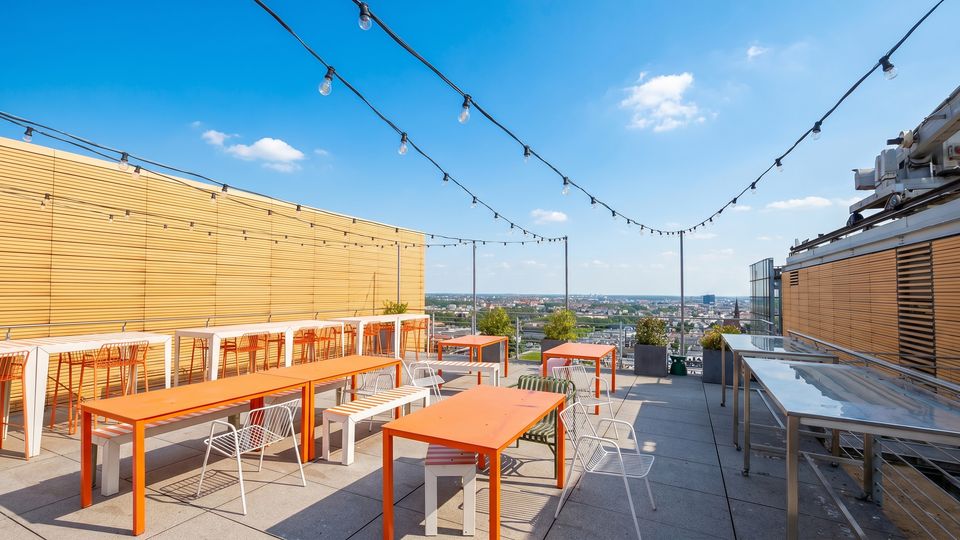 Rooftop terrace with gray paver flooring, orange rectangular tables, and white metal chairs beneath strings of hanging lights. Wooden slat walls frame the space, with planters along the edge and a wide city skyline view in the distance.