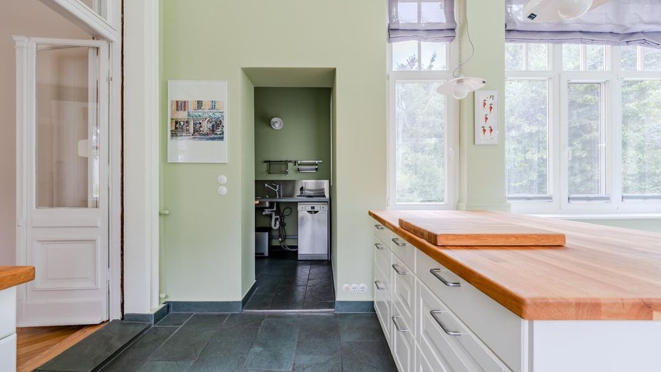 Helle, moderne Küche mit großer Kücheninsel und Arbeitsplatte aus Holz sowie weißen Schubladenschränken auf dunklem Fliesenboden. Große Sprossenfenster lassen viel Tageslicht herein; daneben befindet sich eine Nische mit kleinem Spülbereich.