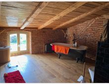 Rustikales Badezimmer in einem Dachgeschoss mit freiliegenden Backsteinwänden und Holzbalkendecke. Im Raum stehen eine freistehende schwarze Badewanne mit orangefarbenem Handtuch, ein kleines Fenster mit Holzrahmen, ein roter Teppich und Holzboden.