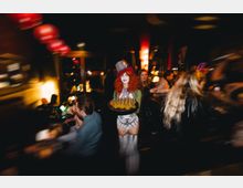 Innenaufnahme einer dunklen, belebten Bar oder eines Nachtclubs mit warmen Lichtquellen, roten Lampions an der Decke und einer langen Theke. Der Raum wirkt eng und ist von zahlreichen Gästen umgeben, mit glänzenden Oberflächen und reflektierenden Fenstern im Hintergrund.