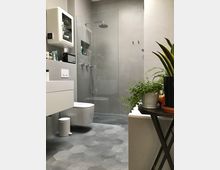 Modernes Badezimmer mit grauen Wänden und sechseckigen Fliesen auf dem Boden. Im Raum befinden sich eine begehbare Dusche mit Glasabtrennung, ein wandhängendes WC, ein weißer Spiegelschrank und Pflanzen auf einem Beistelltisch.