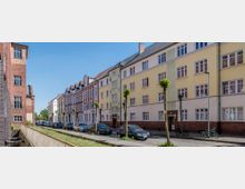 Reihenhausfassaden in einer städtischen Wohnstraße mit gelben und roten Klinkergebäuden, geparkten Autos am Straßenrand und jungen, zurückgeschnittenen Bäumen entlang des Gehwegs. Links im Vordergrund verläuft ein kleiner Kanal mit einem Geländer.