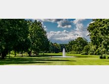 Weitläufiger Park mit einer großen Rasenfläche, umgeben von hohen Bäumen. Im Hintergrund befindet sich ein kleiner Teich mit einer zentralen Wasserfontäne unter einem bewölkten Himmel.