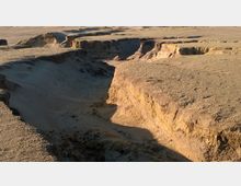 Steinige Wüstenlandschaft mit tiefen und unregelmäßig verlaufenden Schluchten, die von Erosion geformt wurden. Die Oberfläche ist trocken und von sandigen sowie felsigen Bereichen geprägt.