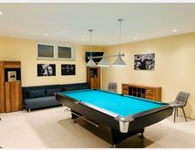 Ein Raum mit einem Billardtisch in der Mitte, beleuchtet von einer Hängelampe mit drei Leuchten. Im Hintergrund stehen ein dunkelgraues Sofa, Holzmöbel und Stühle, und an den beige gestrichenen Wänden hängen Schwarz-Weiß-Bilder.