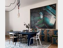 Esszimmer mit einem dunkelblauen Tisch, umgeben von diversen modernen Stühlen. An der weißen Wand hängt ein großes abstraktes Gemälde mit grünen und dunklen Treppenhaus-Motiven. Auf dem Boden liegt ein Teppich, und rechts sind Umzugskartons gestapelt.