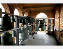 Ein Glockenstuhl mit mehreren großen Metallglocken in einem Turm aus rotbraunem Ziegelmauerwerk. Im Hintergrund sind offene Rundbögen, durch die ein bewaldetes Gebiet sichtbar ist.