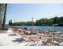 Außenbereich eines Cafés oder Restaurants mit Holztischen und Klappstühlen auf weißem Sand, direkt am Ufer eines Flusses. Im Hintergrund sind Bäume, Wasserfahrzeuge und ein klarer, blauer Himmel zu sehen.
