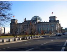 Außenansicht des Reichstagsgebäudes in Berlin mit seiner markanten Glaskuppel auf dem Dach. Im Vordergrund eine Straße mit parkenden und fahrenden Autos sowie einem Gehweg, flankiert von jungen, kahlen Bäumen.