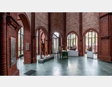 Innenraum einer Kirche mit roten Backsteinwänden und hohen spitzbogigen Fenstern. Im Raum befinden sich Skulpturen, ein Tisch mit liturgischen Gegenständen und Ausstellungsstücke auf Podesten.