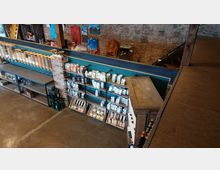 Blick in einen Laden mit Ziegelsteinwänden und industriellem Stil. Regale mit verpackten Lebensmitteln, großen Vorratsbehältern für Schüttgut und Getränkeflaschen dominieren den Raum, während an der Wand Gemälde hängen.