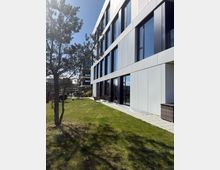 Ein modernes Bürogebäude mit großer Fensterfront und heller Fassadenverkleidung. Im Vordergrund befindet sich eine Rasenfläche mit einzelnen Bäumen, Gartenmöbeln und Gehwegplatten.