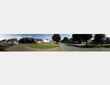 Panoramaaufnahme eines ländlichen Dorfplatzes mit einer breiten Straßenkreuzung, gepflegten Grünflächen und Bäumen. Umgeben von kleinen Wohnhäusern mit teils weißen, teils roten Fassaden und Satteldächern.