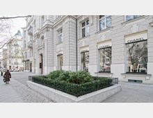 Straßenszene mit einem eleganten Gebäude aus hellem Stein, in dem mehrere Chanel-Schaufenster eingelassen sind. Vor dem Gebäude befindet sich ein gepflegtes, von einem niedrigen Geländer umgebenes Beet, während Bürgersteige und Bäume die Umgebung prägen.