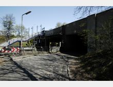 Ansicht eines Bahnhofsbereichs mit einer blauen Treppe, die zu einer Bahnsteigüberführung führt, neben einer Unterführung aus Beton. Eine Kopfsteinpflasterstraße, ein Straßenschild und ein Fahrradständer befinden sich im Vordergrund, umgeben von Bäumen und Sträuchern.