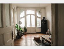 Helles Wohnzimmer mit Holzboden, einem großen bogenförmigen Fenster mit weißen Holzrahmen, mehreren Zimmerpflanzen, einem niedrigen Tisch auf Rollen, einem runden Hocker und einem schwarzen Schrank. Im Hintergrund sind Bücherstapel und eine Pendelleuchte sichtbar.