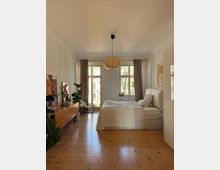 Bright, minimalist bedroom with a wooden floor, a double bed with white bedding, and a woven pendant light centered on the ceiling. Two large windows and a glass door with gray curtains face a balcony, with a long wooden sideboard, plants, and framed artwork along the left wall.