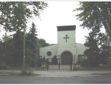 Kirchengebäude mit schlichtem, weißem Putz und einem zentralen Turm mit schwarzem Kreuz an der Fassade. Vor dem Gebäude befinden sich ein Zaun, ein Tor und einige Bäume, die das Grundstück umgeben.