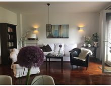 Wohnzimmer mit Holzfußboden, einer weißen Couch und einem schwarzen Sessel. Eine Bücherwand, ein Kamin mit Dekorationen und ein Wandbild schmücken den Raum; Zimmerpflanzen und ein großer Blütenstand auf einem Tisch im Vordergrund ergänzen das helle und modern eingerichtete Ambiente.