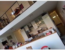 Wohnraum mit offener Küche und Zwischengeschoss. Im unteren Bereich befinden sich eine Holzarbeitsplatte mit Küchenschränken, ein Waschbecken und Küchengeräte; darüber ein Geländer im Zwischengeschoss mit Büchern und Möbeln.