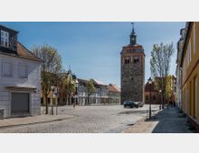 Historischer Marktplatz mit gepflasterter Fläche, umgeben von bunten Gebäuden mit Satteldächern; im Hintergrund steht ein massiver Steinturm mit einer Uhr und rotem Backsteingiebel, flankiert von Bäumen.