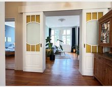 Innenraum eines Hauses mit Holzboden und weißen Flügeltüren mit Glaseinsätzen in Gelb und Weiß. Im Hintergrund ein Wohnzimmer mit großen Fenstern, Gardinen und Zimmerpflanzen; links ein Schlafzimmer mit blauer Wand und einem Holzbett.