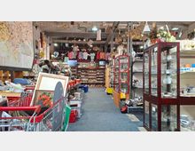 Innenansicht eines großen Secondhand- bzw. Trödelladens in einer Industriehalle mit hohen Decken, vielen hängenden Lampen und dicht gefüllten Regalen. Ein breiter Gang führt zwischen Vitrinen mit Glaswaren, Kisten, Einkaufswagen und ausgestellten Gegenständen bis zu einer Rückwand mit weiteren Regalen und aufgehängter Kleidung.