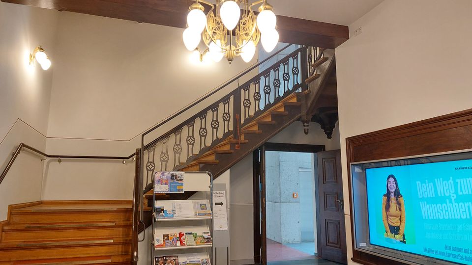 Ein Eingangsbereich mit einer Holztreppe mit verzierten Metallgeländern, einer hängenden Kronleuchterbeleuchtung und einer Wandhalterung mit Prospekten. Rechts befindet sich ein Flachbildschirm mit einer Werbeanzeige, daneben eine offene Holztür.