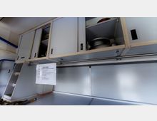 Innenansicht eines Küchenwagens mit weißen, verschließbaren Oberschränken, teilweise geöffnet und gefüllt mit Küchenutensilien. Darunter befindet sich eine Arbeitsfläche, an der ein Zettel mit Informationen befestigt ist.