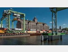 Historisches Lagerhaus aus rotem Backstein mit der Aufschrift „Behala Westhafen“, im Vordergrund Containerkräne, gestapelte Frachtcontainer und ein Wasserkanal. Die Umgebung zeigt Hafeneinrichtungen und ein Gebäude im Hintergrund.