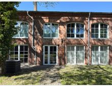 Zweistöckiges Gebäude aus rotem Backstein mit großen, weiß umrahmten Fenstern und Türen in symmetrischer Anordnung. Im Vordergrund eine grüne Rasenfläche und ein gepflasterter Weg, teils im Schatten von Bäumen.