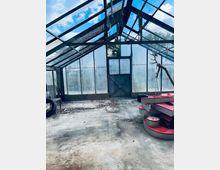 Ein leeres, verglastes Gewächshaus mit Metallrahmen und bläulichen Dachscheiben, das einen verwitterten Betonboden aufweist. Im Vordergrund liegen rote, kreisförmige Objekte und ein Geweih; im Hintergrund sind Doppeltüren und angelaufene Seitenfenster erkennbar.