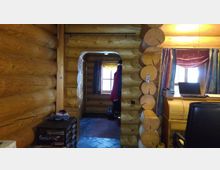 Innenraum einer Blockhütte mit sichtbaren Holzstämmen als Wände. Ein Durchgang führt zu einem Fenster mit Vorhängen, daneben ein Schreibtisch mit Laptop und im Vordergrund ein schwarzer Bürostuhl.