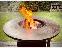 Runder, metallener Feuerkorb mit breiter Grillplatte und zentraler Öffnung, in der Holzscheite mit sichtbaren Flammen brennen. Dahinter ein Garten mit Rasenfläche, dichter Heckenbepflanzung und einem Holzlagerregal am Rand.