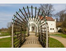 Eine mit Holzlatten und Metallbögen gestaltete Pergola führt zu einem weißen Gebäude mit roten Ziegeln, das von Rasenflächen, Bäumen und einem gepflasterten Weg umgeben ist. Im Hintergrund sind kahle Bäume und ein klarer Himmel zu sehen.