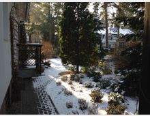 Ein verschneiter Garten mit einem gepflasterten Weg, der entlang eines Hauses verläuft. Im Garten stehen Bäume und Sträucher, teils bedeckt mit Schnee, im Hintergrund sind weitere Häuser und Wald erkennbar.