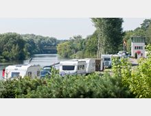 Wohnmobilstellplatz am Ufer eines Flusses mit dichtem Baumbewuchs im Hintergrund; im Vordergrund stehen Wohnmobile und Wohnwagen entlang einer schmalen Straße, daneben ein kleines Gebäude mit Fahne und grünem Dach.