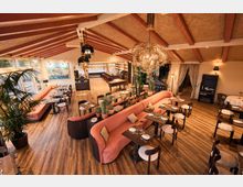 Spacious restaurant dining room with wood floors, exposed ceiling beams, and large woven pendant lights. Coral upholstered banquette seating surrounds set tables, with potted plants, tall windows along one side, and a bar area in the back.