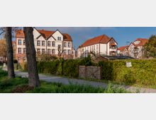 Wohnsiedlung mit historischen Gebäuden aus rotem Backstein und weißen Fassaden mit roten Dächern, umgeben von einer dichten Hecke und einem Gehweg im Vordergrund, gesäumt von großen Bäumen.