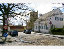 Straßenszene in einer historischen Altstadt mit Kopfsteinpflaster, einem weißen Gebäude mit grünen Fensterläden und der Beschilderung „Chopin – Restaurant“, im Hintergrund eine Kirche mit einem rechteckigen Turm. Parkende Autos und Verkehrszeichen, wie eine Geschwindigkeitsbegrenzung auf 30 km/h, sind zu sehen, umgeben von kahlen Bäumen.