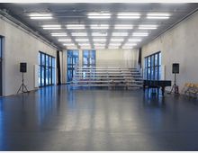 Großer, moderner Proberaum mit grauem Bodenbelag, hohen Decken und großen, bodentiefen Fenstern an den Seitenwänden. Der Raum enthält mobile Tribünen, einen schwarzen Flügel, Stuhlreihen und zwei Lautsprecher auf Stativen.