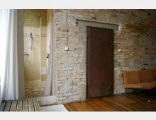 Raum mit offenem Zugang zu einer Dusche mit beigefarbenen Fliesen und Mosaikboden, rechts eine gemauerte Ziegelwand mit einer schweren, dunklen Stahltür sowie einer Wandhalterung; Holzboden und ein orangefarbenes Sofa im Vordergrund.