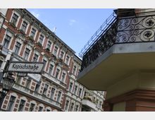 Straßenszene mit Altbau-Fassaden in rotem Klinker und weißem Stuck, einem dekorativen Balkon mit filigranem Gittergeländer im Vordergrund sowie einem Straßenschild mit der Aufschrift "Kopischstraße". Der Himmel ist klar und blau.
